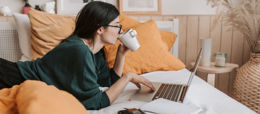 Woman lying on bed sipping coffee and working on a laptop