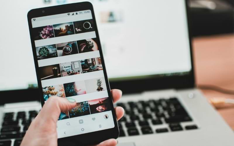 A hand holding a phone that has a collection of photos on it. In the background is a laptop, although the screen is blurred. 