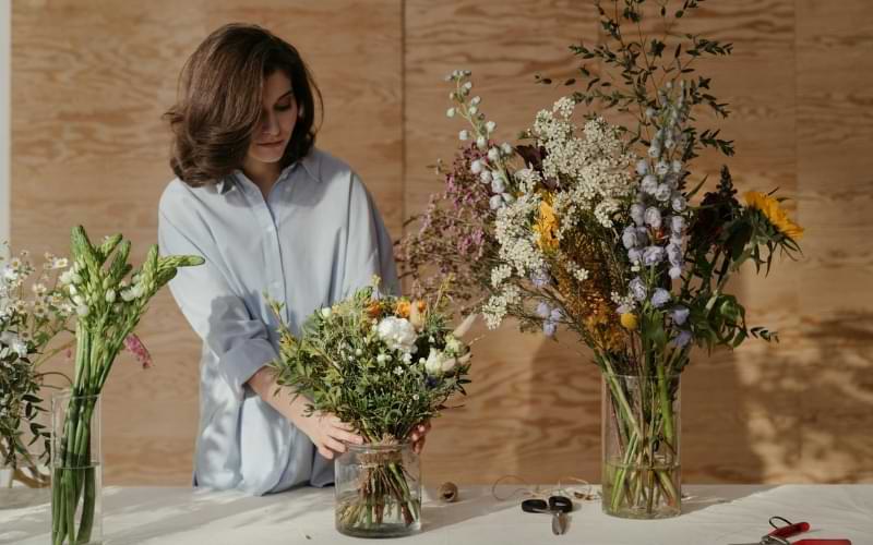 A woman in a blue shirt making spring bouquets. There are three bouquets in three glass vases. 